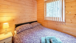 Una habitación de cabaña de madera con una cama, una mesita de noche, una lámpara y una ventana con persianas.