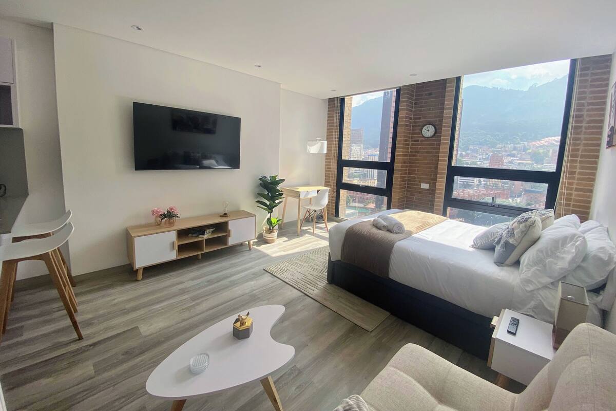 A modern hotel room with a large bed, a TV mounted on the wall, a desk, and a view of the city through large windows.