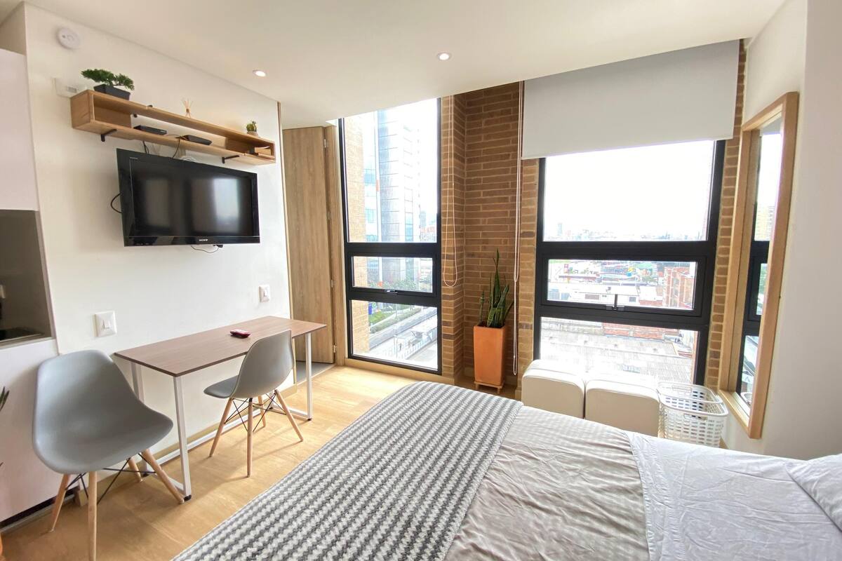 A modern hotel room with a large bed, a desk with a chair, a flat-screen TV, and large windows offering a city view.