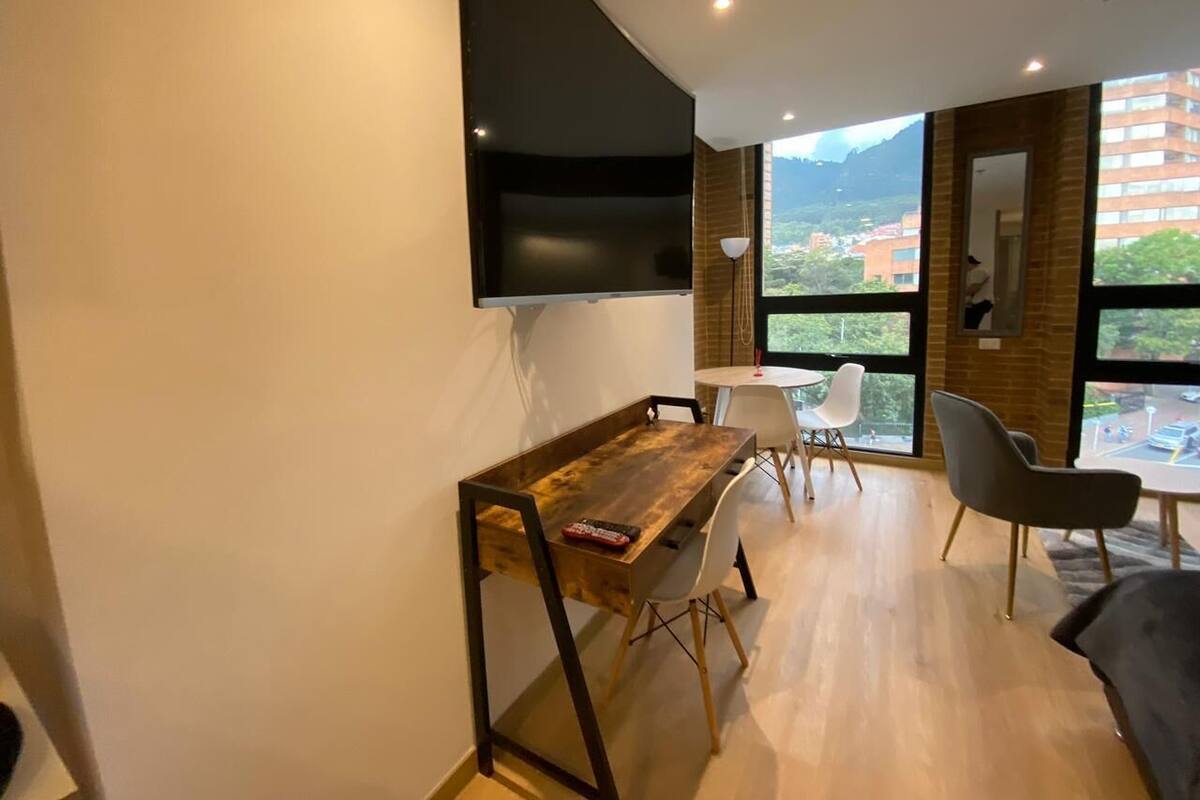 A modern living room with a wooden coffee table, white chairs, a flat-screen TV mounted on the wall, and large windows offering a view of the cityscape.