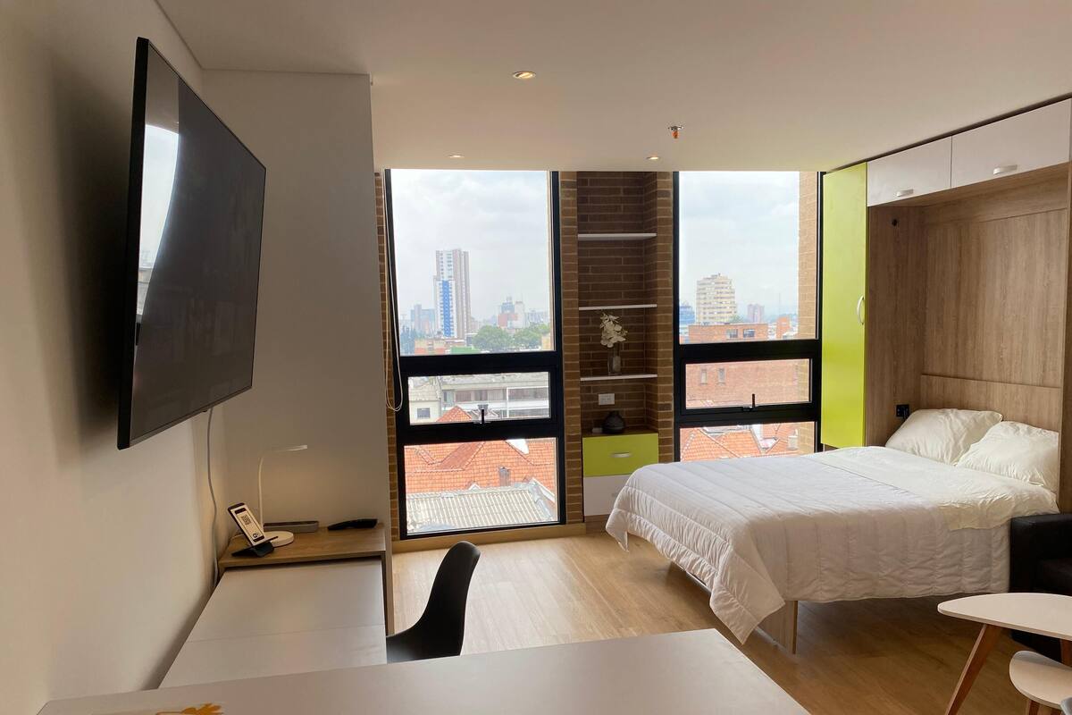 A modern bedroom with a bed, a desk, a chair, a TV, and a large window with a city view.