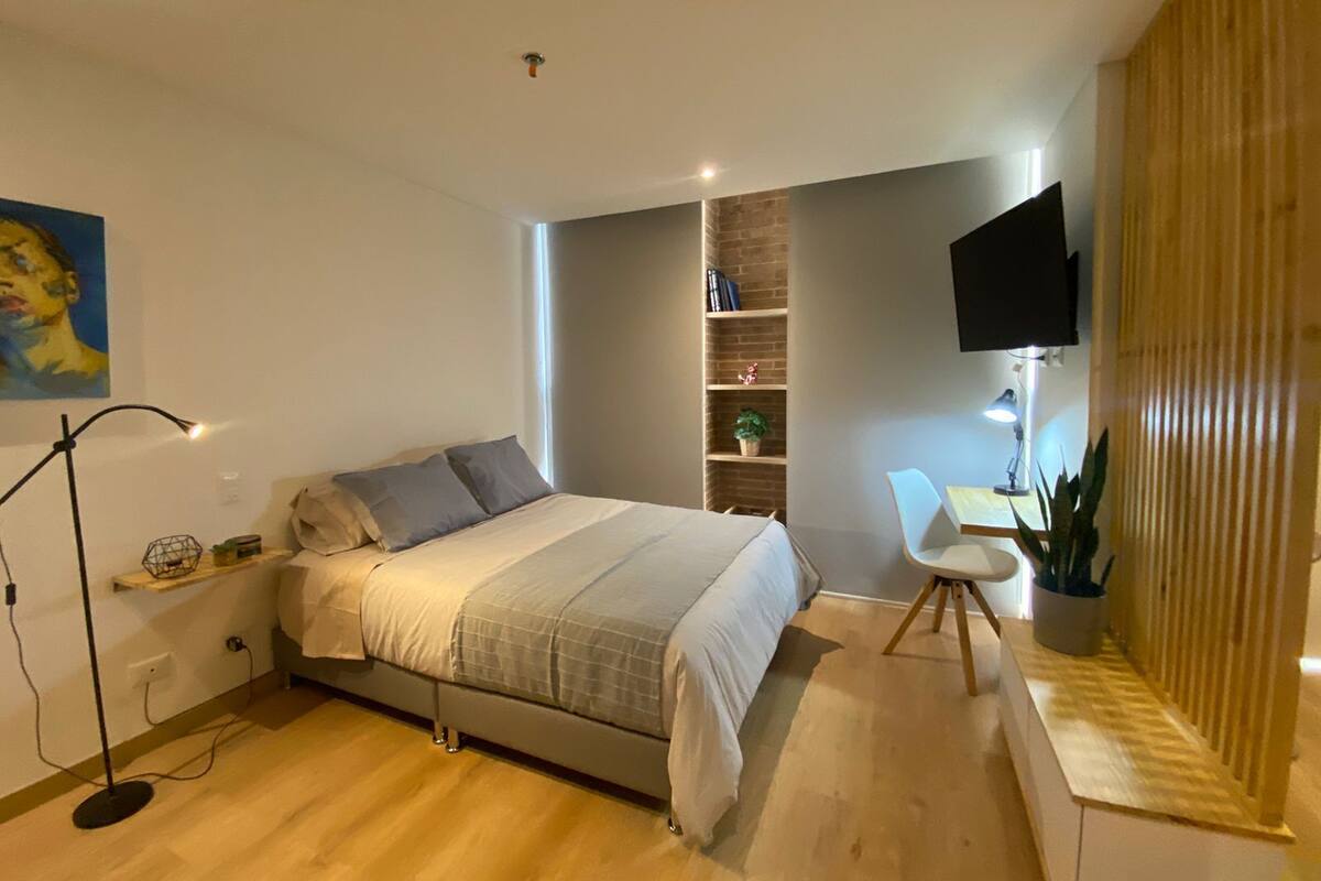 A modern bedroom with a bed, bedside table, desk, chair, and a TV mounted on the wall.