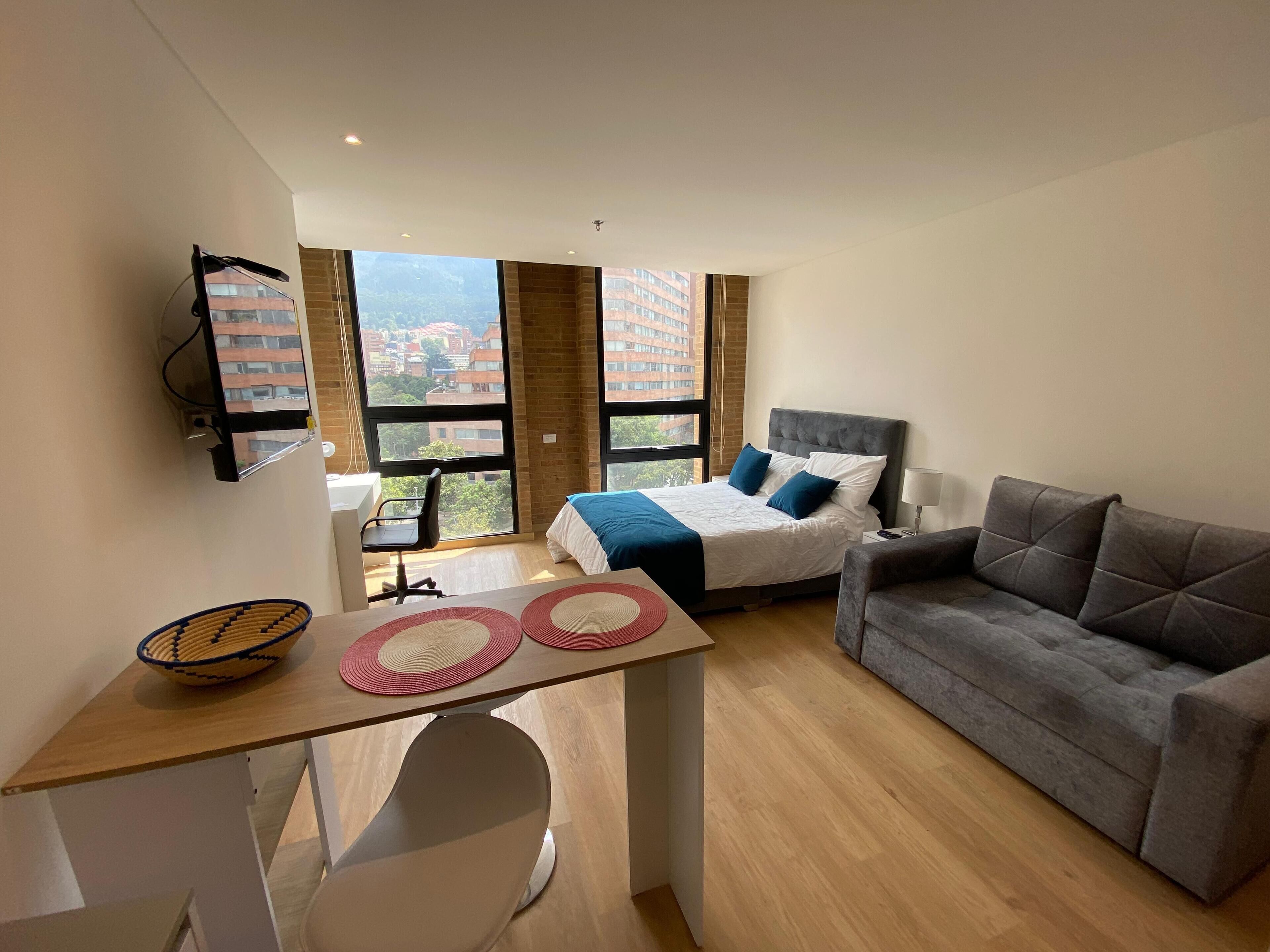 Een moderne hotelkamer met een bed, een grijze bank, een bureau met stoel, en grote ramen met uitzicht op de stad.