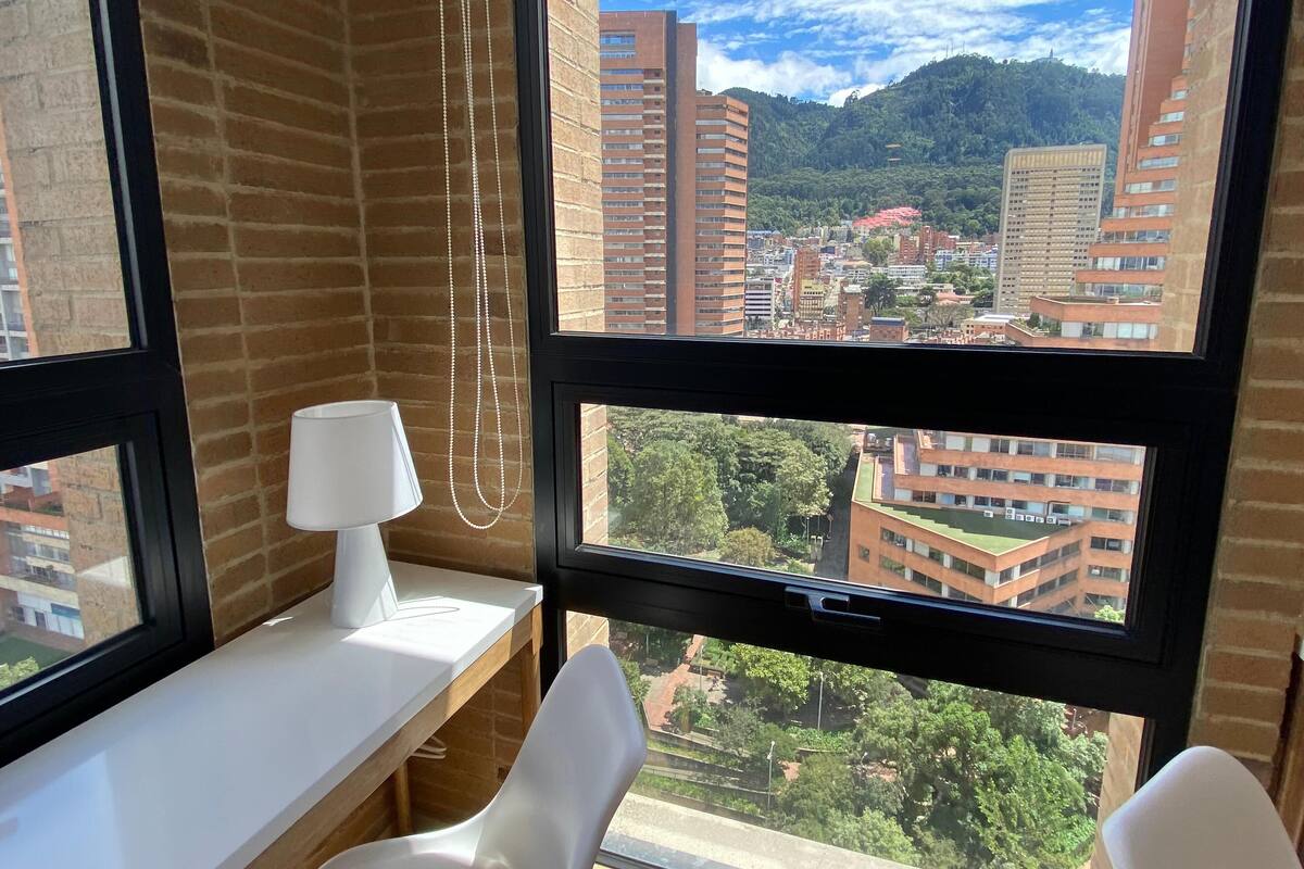 A room with a desk, a chair, a lamp, and a view of a cityscape with buildings and greenery.
