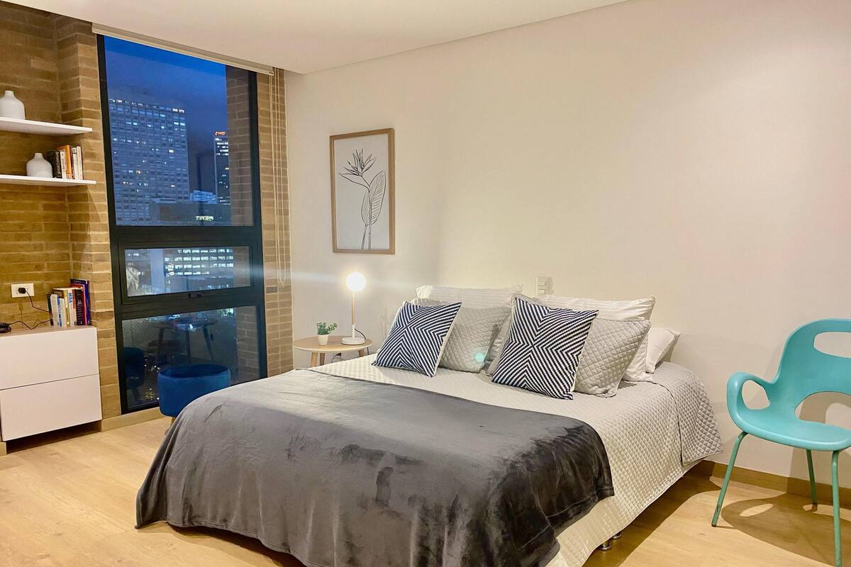 A modern bedroom with a large bed, a view of the city through a window, a small table with a lamp, and a chair.