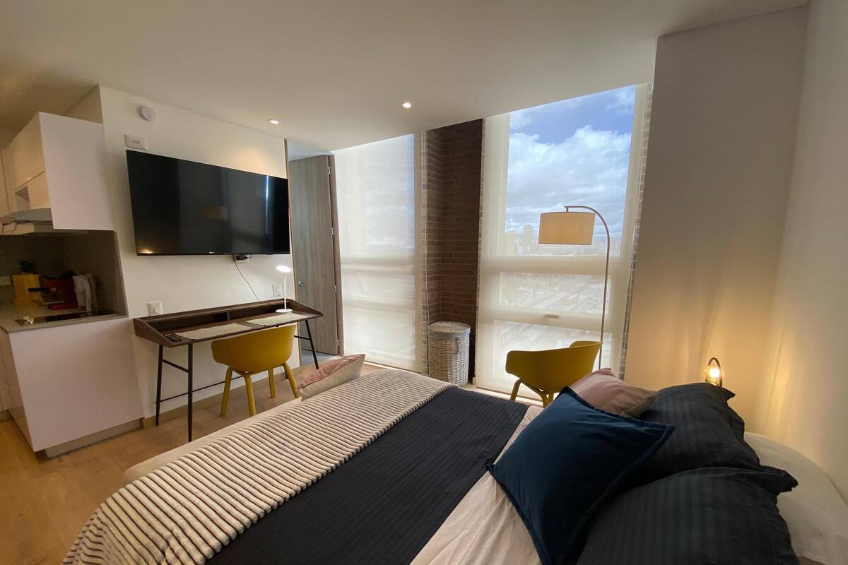 A modern hotel room with a bed, a desk, a chair, a TV, and a large window with a city view.