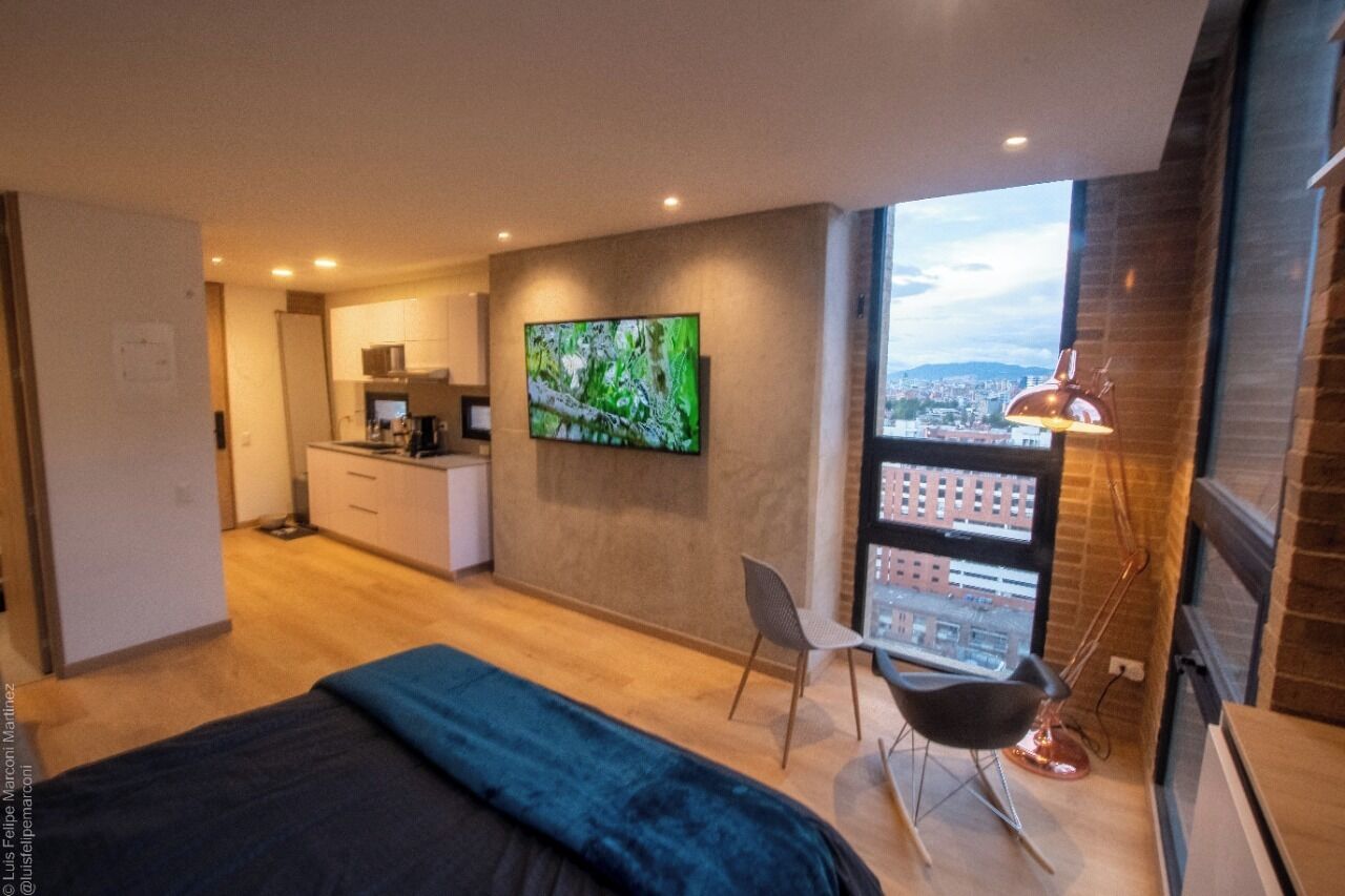 Ein modernes Hotelzimmer mit einem großen Bett, einem an der Wand montierten Fernseher, einem Essbereich und Blick auf die Stadt durch große Fenster.