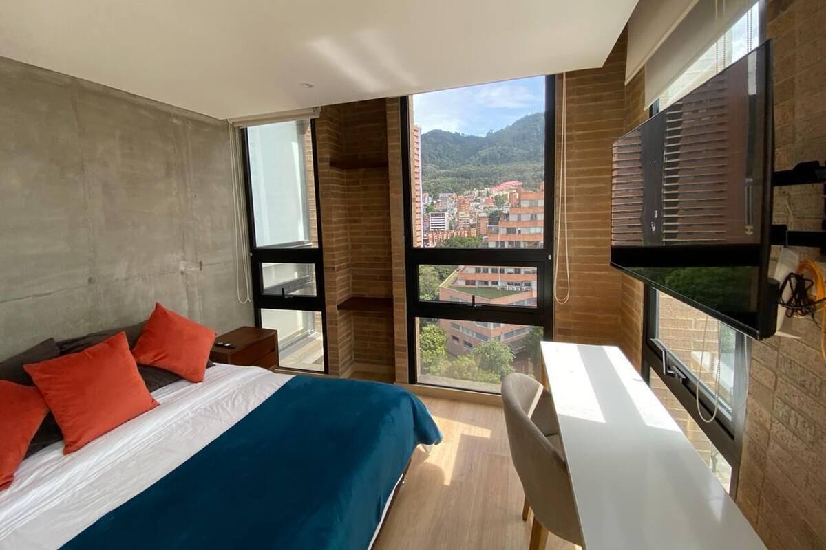 A modern bedroom with a large bed, a desk, and a view of a cityscape through large windows.