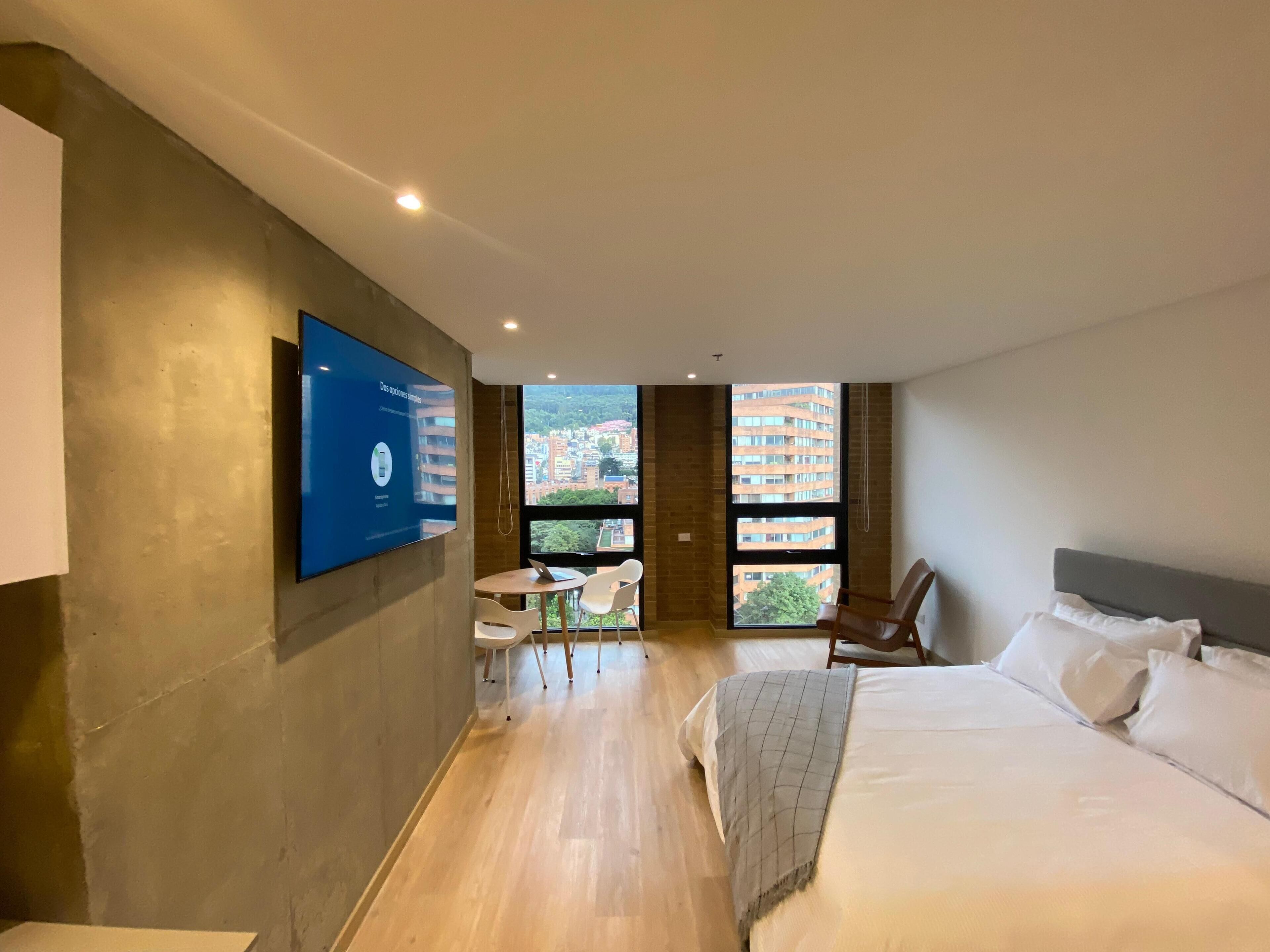 A modern hotel room with a large bed, a desk with chairs, a TV mounted on the wall, and large windows offering a city view.