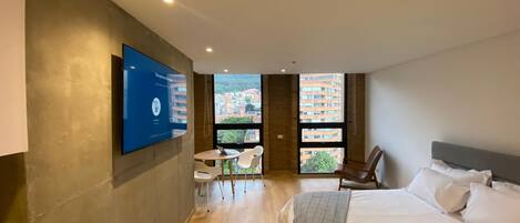 A modern hotel room with a large bed, a desk with chairs, a TV mounted on the wall, and large windows offering a city view.