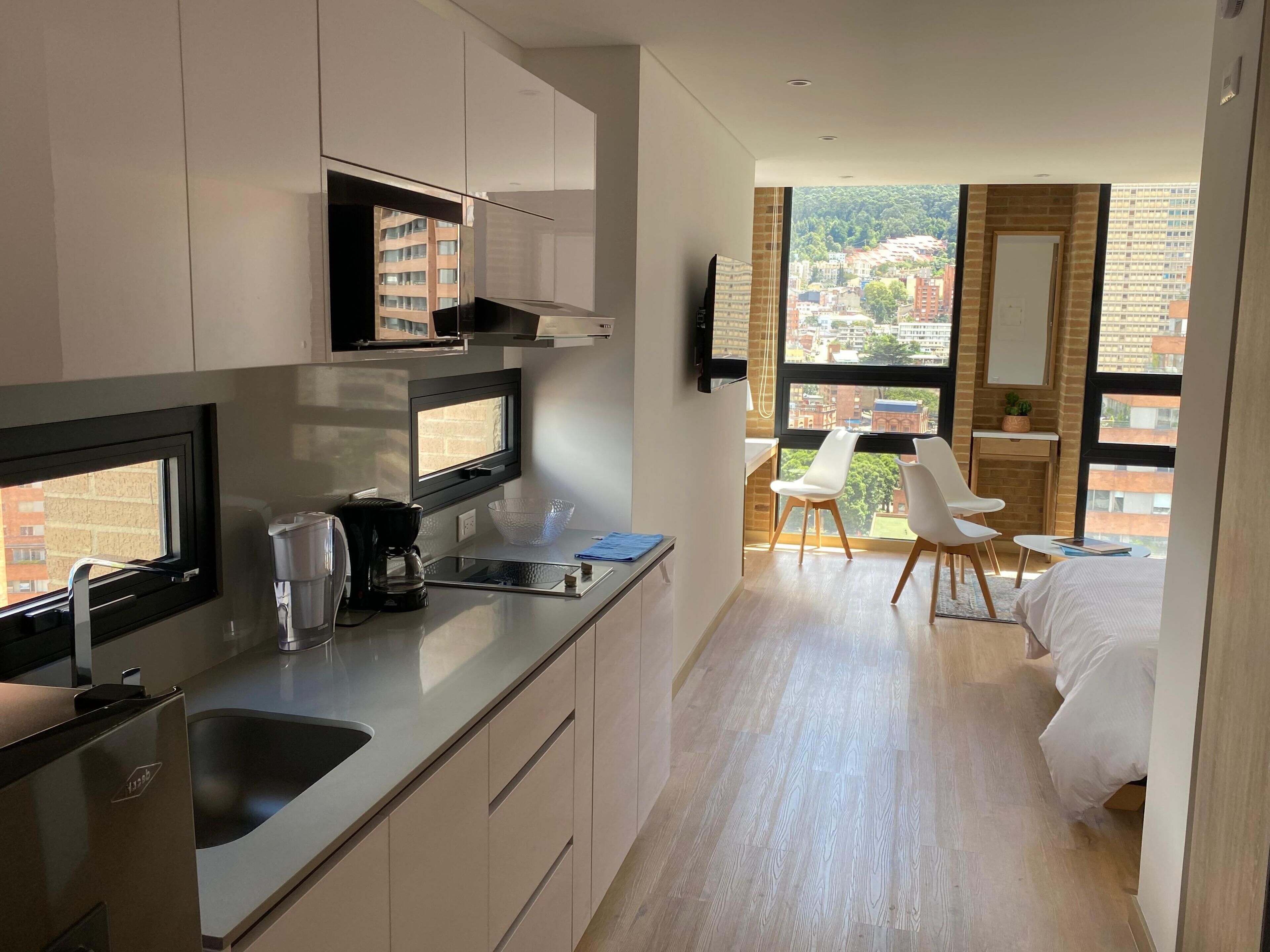 A modern hotel room with a kitchenette, a bed, a dining area with a view, and a large window.