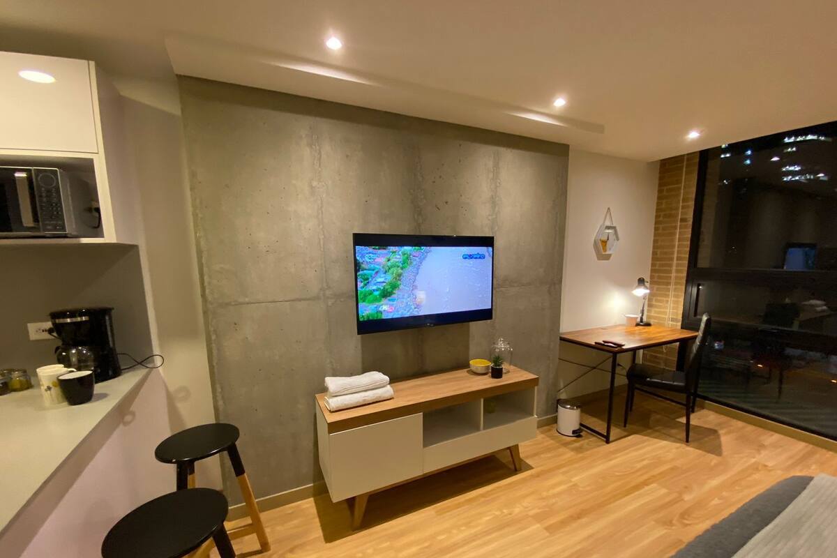 A modern hotel room with a flat-screen TV mounted on the wall, a wooden desk, a small seating area with stools, and a large window offering a view of the city.
