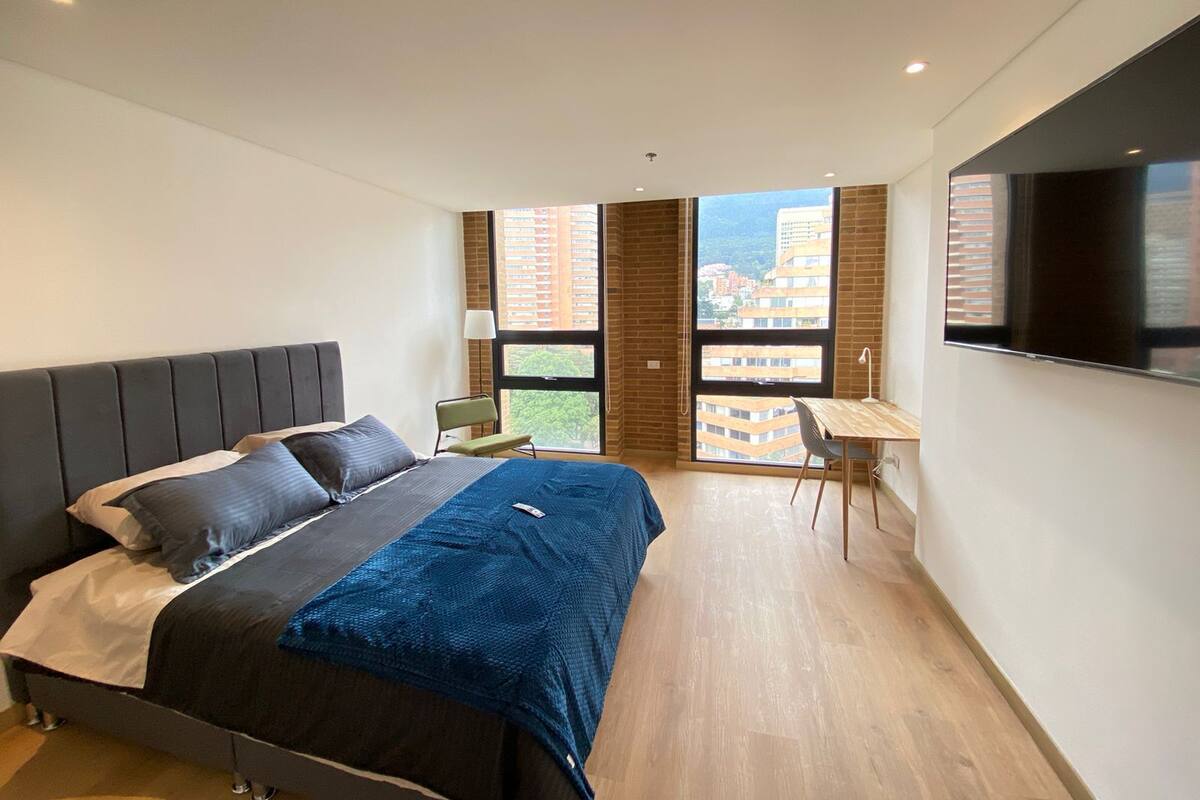 A modern bedroom with a bed, a desk, and a large window offering a city view.