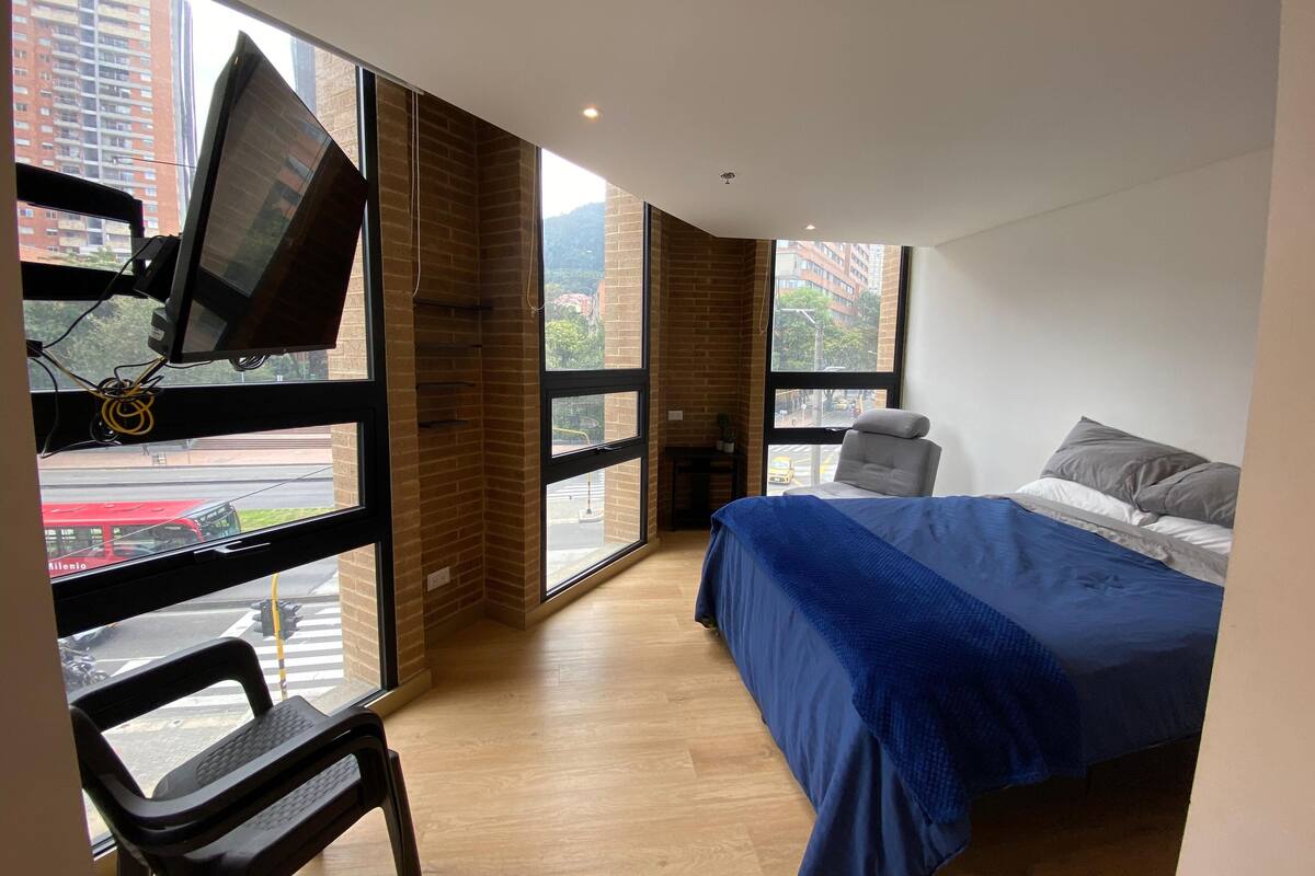 A bedroom with a bed, a chair, a TV mounted on the wall, and large windows with a view of buildings and a street.