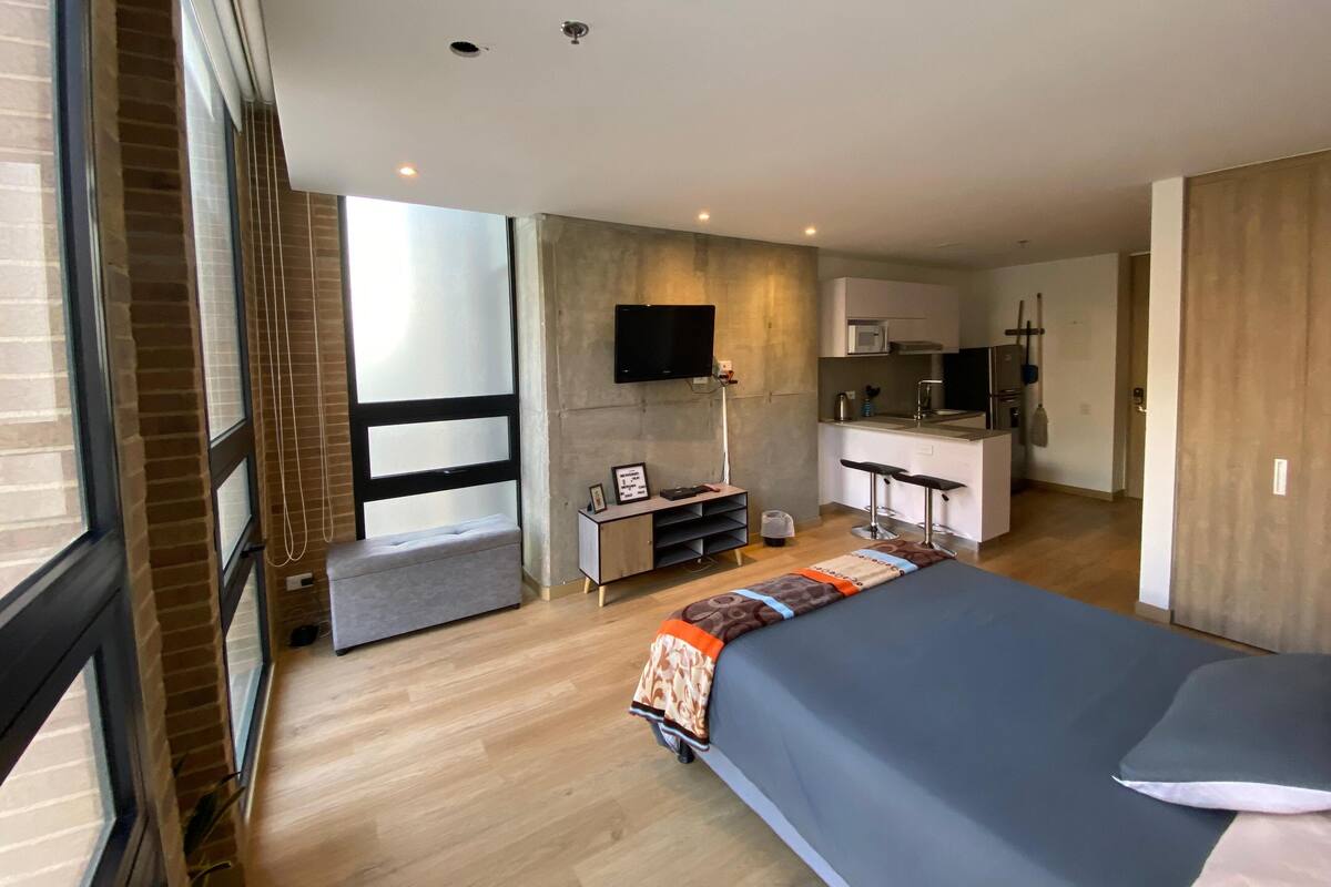 A modern hotel room with a large bed, a desk with a chair, a flat-screen TV mounted on the wall, a kitchenette with a microwave and sink, and a large window with blinds.