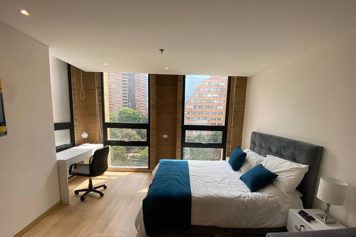 A modern hotel room with a large bed, a desk, and a chair. There are two windows providing a view of tall buildings and trees.