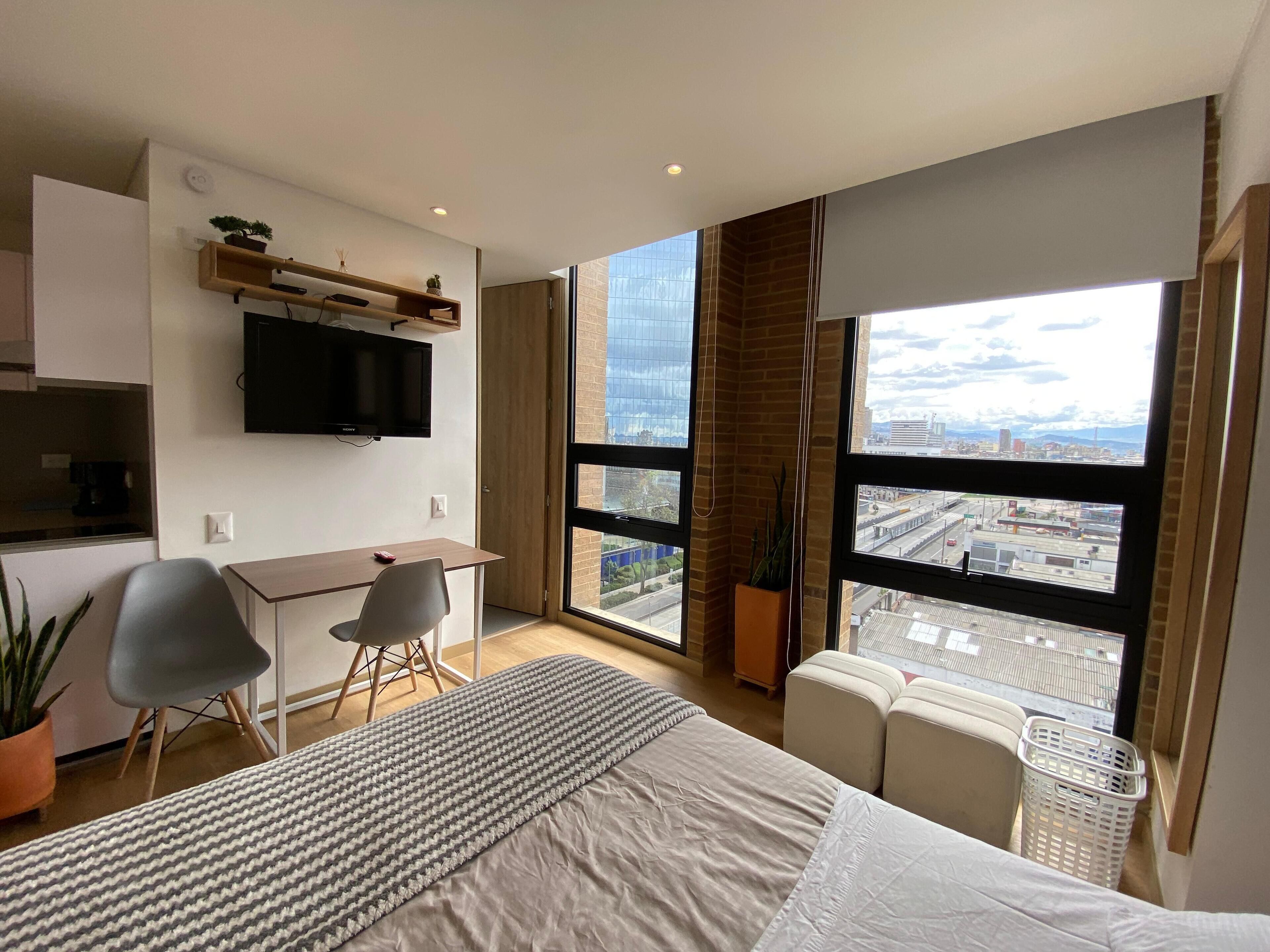 A modern hotel room with a large bed, a desk, a chair, a TV, and a view of the city through large windows.