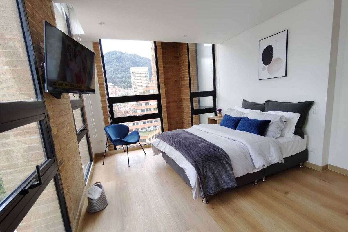 A modern bedroom with a large bed, a TV mounted on the wall, and a view of the city through large windows.
