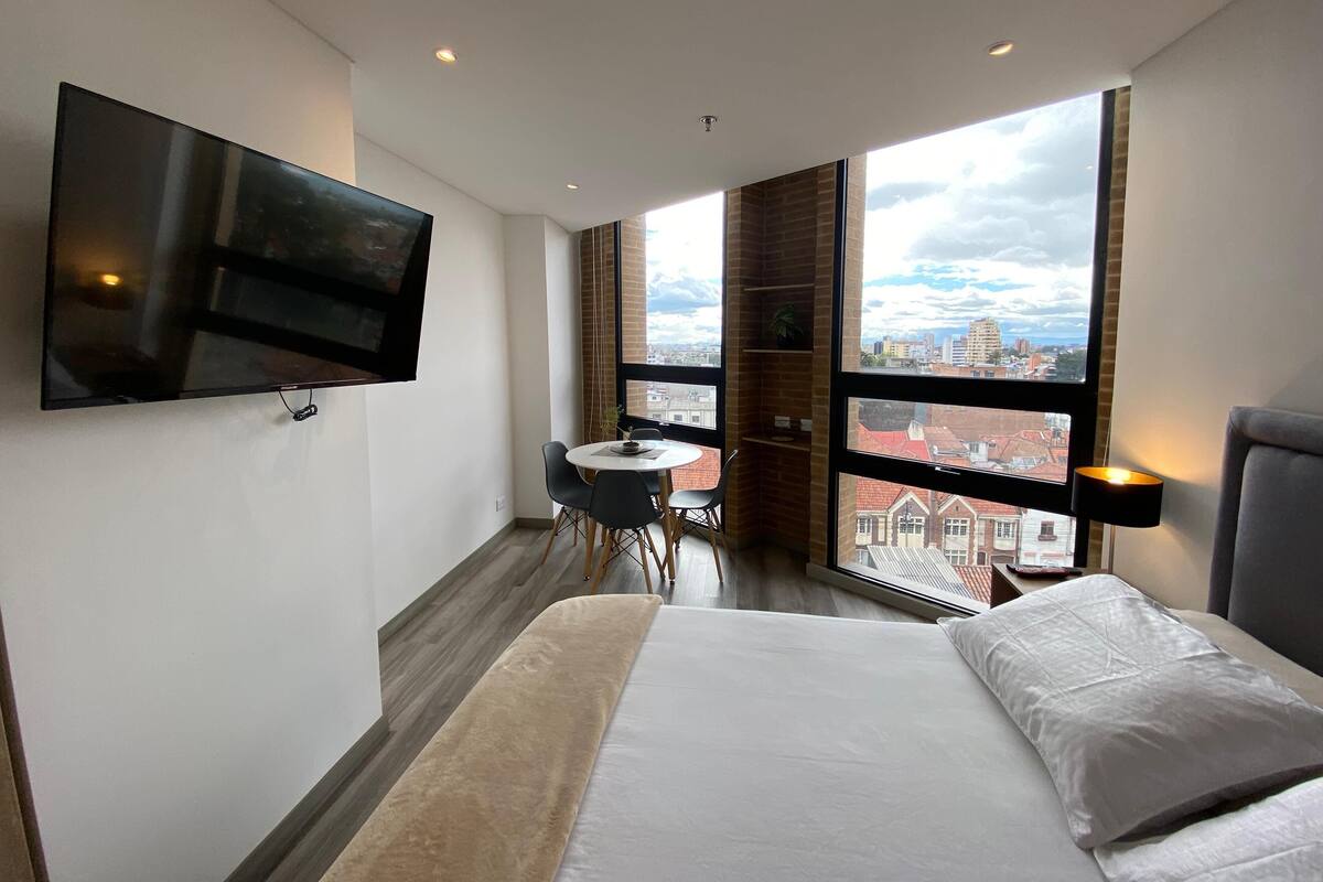A modern hotel room with a large bed, a TV mounted on the wall, a small dining table, and a view of the city through large windows.