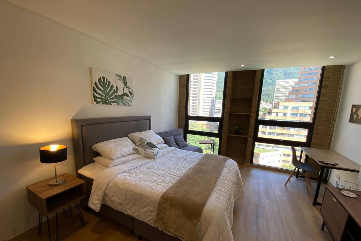 A modern bedroom with a large bed, a desk, and a view of buildings through large windows.