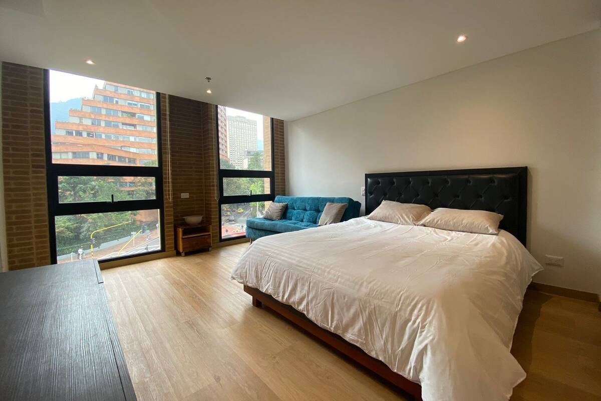 A modern bedroom with a large bed, a blue sofa, and a view of a cityscape through large windows.