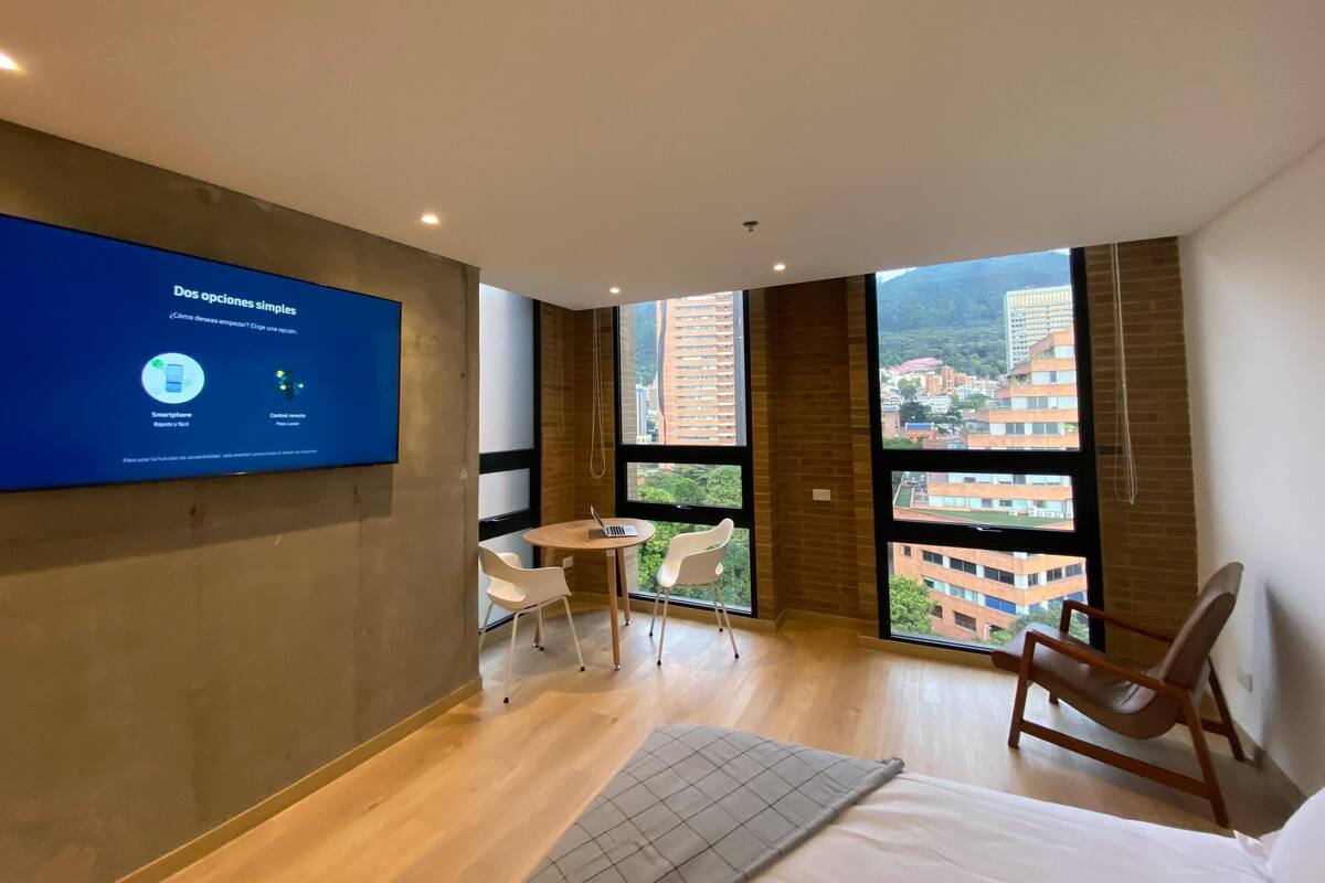 A modern hotel room with a large TV displaying a welcome message, a bed, a desk with a laptop, a chair, and large windows offering a city view.