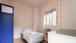Une petite chambre avec un lit simple, un bureau et une chaise, une fenêtre et une porte bleue.