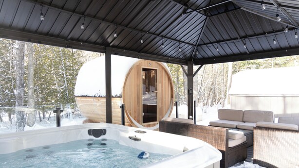 A hot tub area with a wooden sauna, outdoor seating, and a covered patio. - Winter Water Woodlands - Hot Tub & Sauna Near Boyne Mountain & Lake Charlevoix (Boyne City)