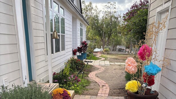 Ein Weg führt zu einem Haus mit Ziegelbordurierung, umgeben von verschiedenen Kübelpflanzen und Blumen.