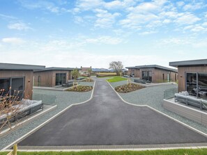 A modern building complex with a central paved walkway, outdoor seating areas, and a view of the surrounding landscape. - Lligwy Lodge 3 - UK42793 (Llangefni)