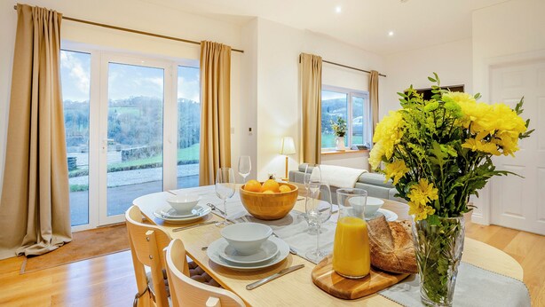 A dining area with a table set for two, a bowl of fruit, a glass of orange juice, and a vase of flowers.