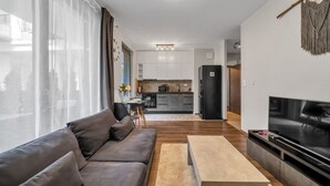 A modern living room with a sofa, coffee table, and TV. There is a kitchen in the background with cabinets and appliances. - Kasprzaka 29C - Terrace, Workspace, EXPO - by Rentujemy (Warszawa)