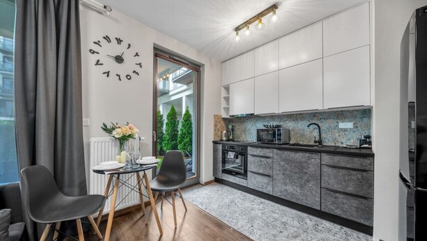 A modern kitchen with white cabinets, a built-in oven, and a microwave. There is a dining area with two chairs and a table, and a clock on the wall. - Kasprzaka 29C - Terrace, Workspace, EXPO - by Rentujemy (Warszawa)