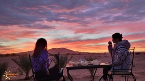 Due persone sedute a un tavolo con vista sul tramonto, paesaggio desertico e un cartello per Harmony Luxury Camp.