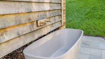 A bathtub outside a wooden house with a grassy lawn in the background.