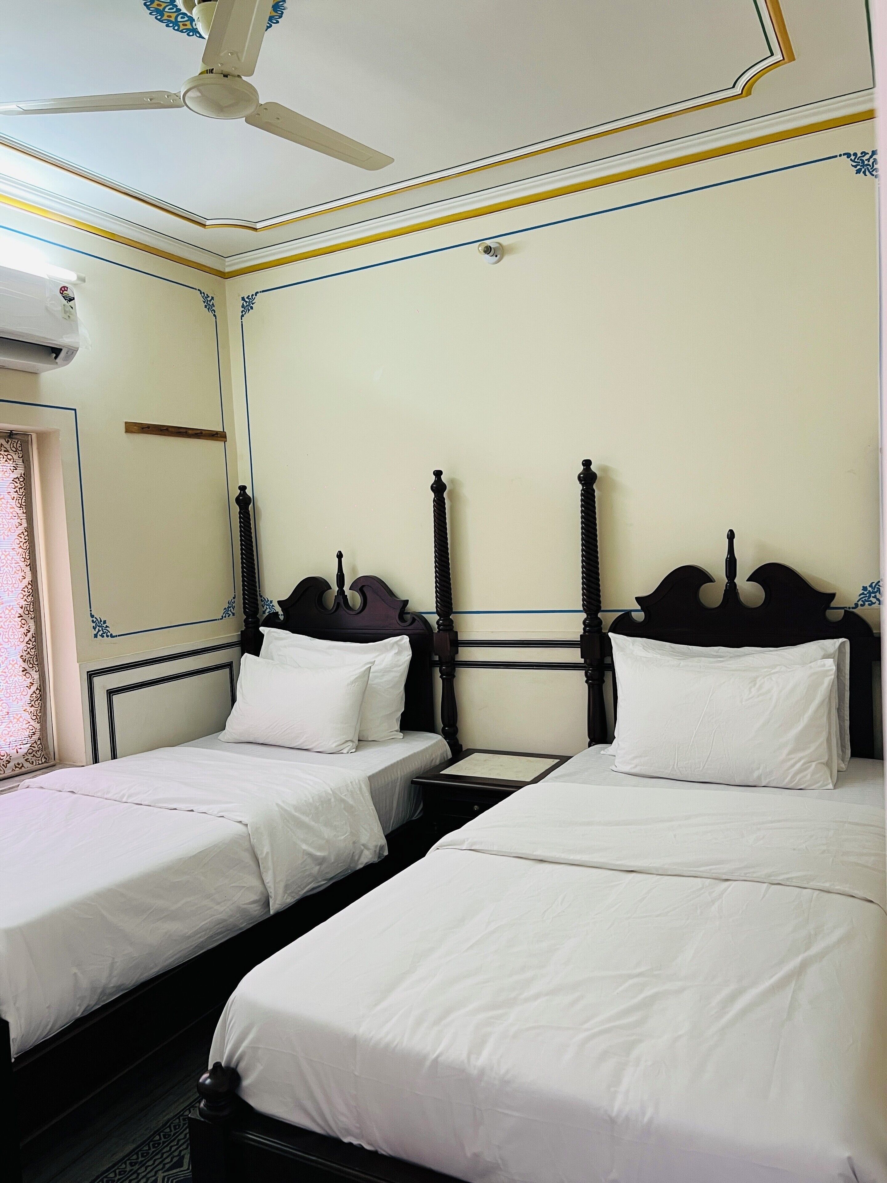 A hotel room with two single beds, a ceiling fan, and a window with patterned curtains.