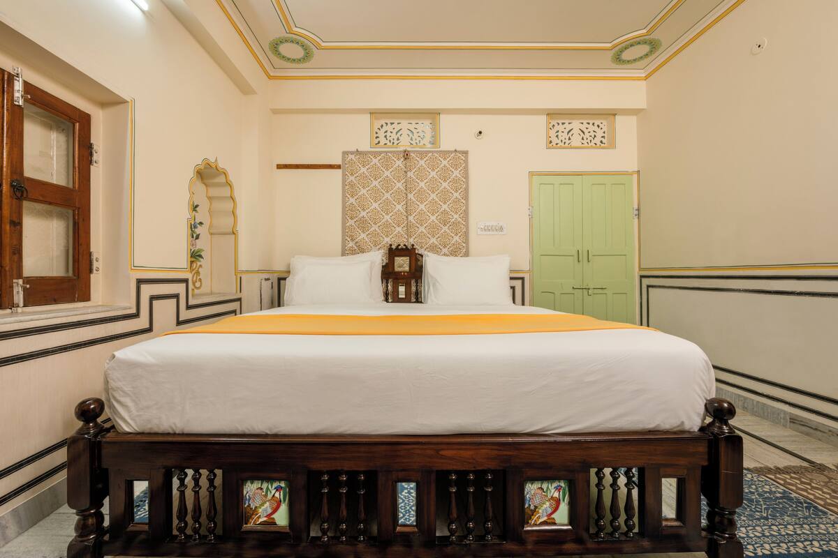 Un lit bien fait, recouvert d’une couvre-lit blanche et orange, avec une tête de lit en bois ornée de motifs complexes, et une porte verte en arrière-plan.