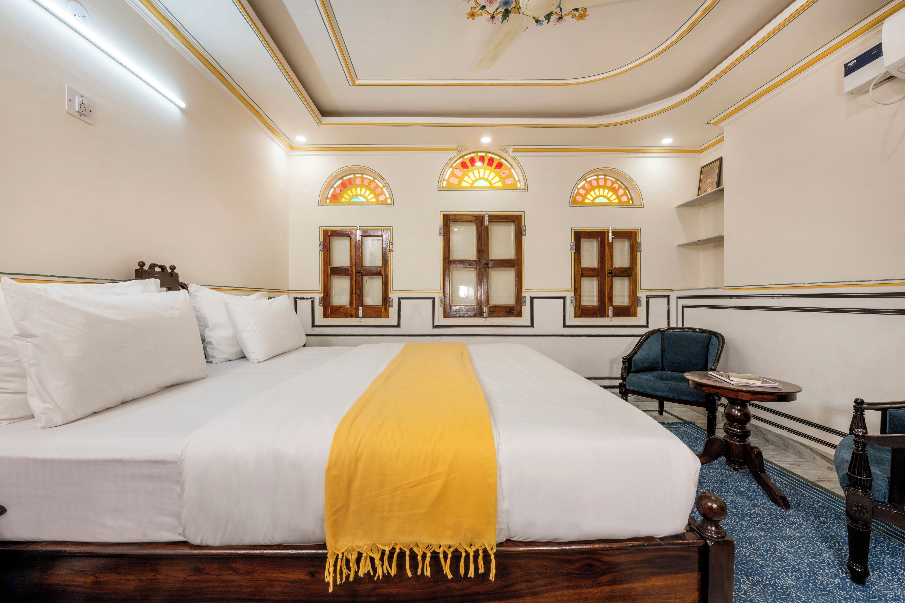 Habitación de hotel con una cama grande, un sillón azul, una mesita redonda y un techo decorativo.