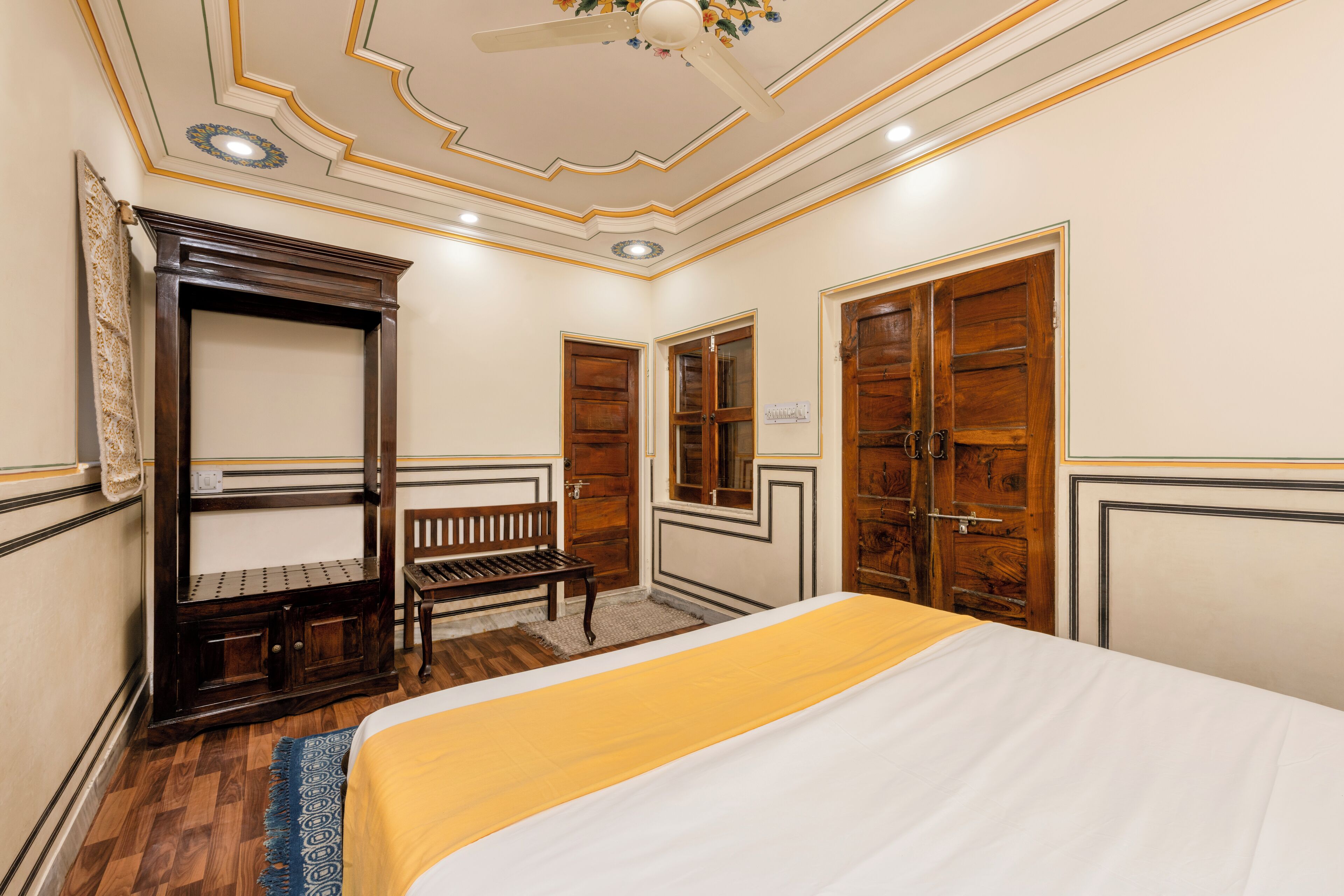 A bedroom with a bed, wooden wardrobe, bench, and a ceiling fan.