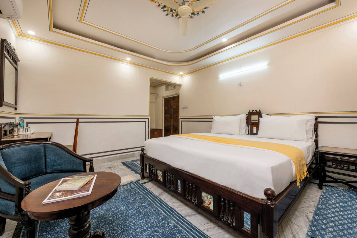 Une chambre d’hôtel avec un grand lit, une table de chevet en bois, un fauteuil bleu, une petite table ronde avec un livre et un tapis bleu à motifs.