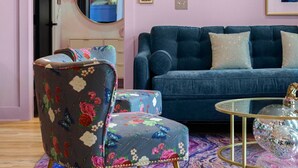 A room with a blue sofa, a floral armchair, a round mirror, and a wall adorned with framed photos and a clock.