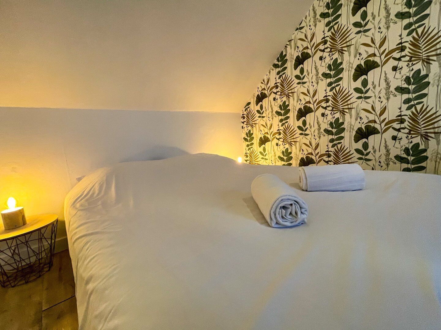 A neatly made bed with a rolled white towel, a bedside lamp, and a wall with a leafy pattern.