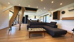 A spacious living area with a wooden staircase, a dark sectional sofa, a glass coffee table, and a kitchen in the background. - house Porzh Loaz. (Plouha)