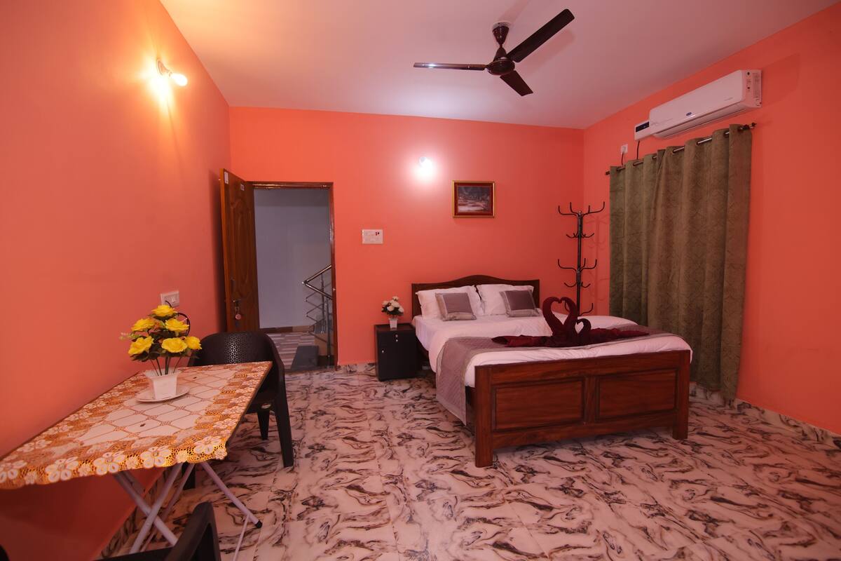 Une chambre à coucher avec un lit, une table sur laquelle est posé un vase de fleurs, un ventilateur de plafond et un climatiseur.