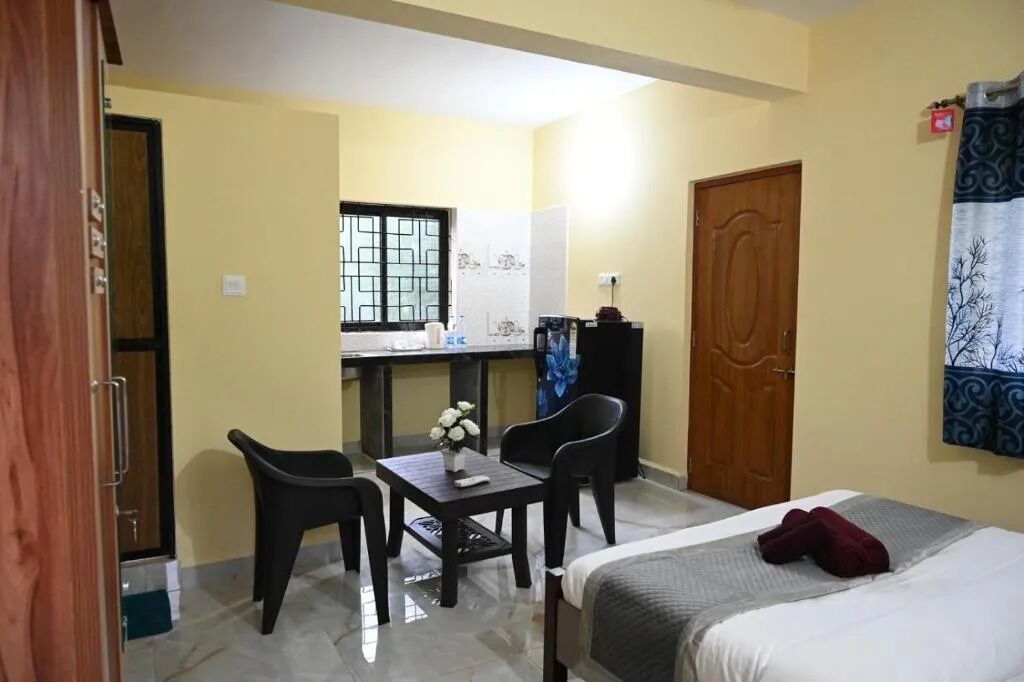 Una habitación pequeña y funcional con cama, escritorio con silla, una mesita con un florero y una ventana con rejas.