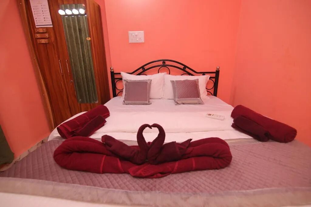 A neatly made bed with a heart-shaped towel arrangement, a wooden door with a mirror, and a simple wall-mounted light switch.