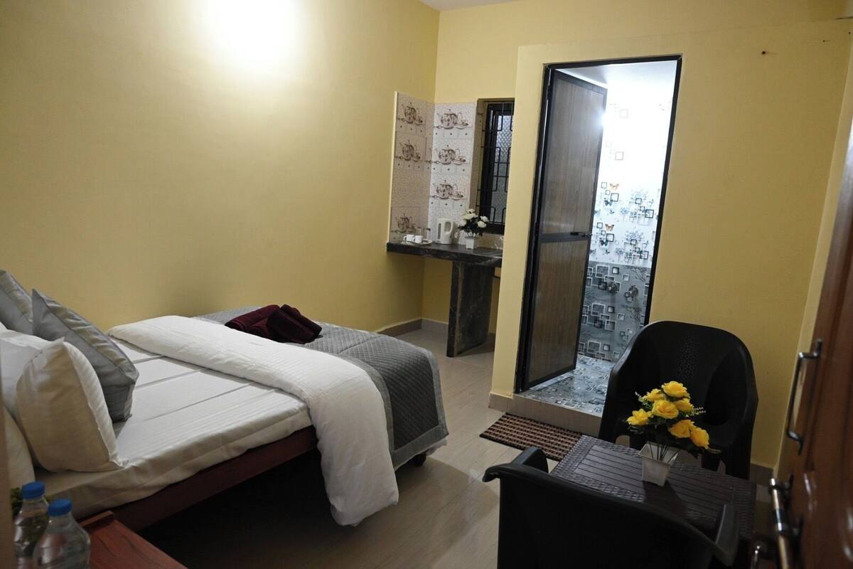 Une chambre d’hôtel avec un lit, un bureau et une chaise, une petite table avec un vase de fleurs, et une vue sur un balcon à travers une porte vitrée.