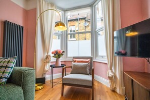A cozy room with a large window, a chair, a small table with a vase of flowers, and a television on a stand.