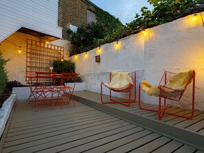 An outdoor patio with red metal chairs and a table, a white building with a window, and a wall with string lights. - Cute Cosy Cottage | Ramsgate Historic Area | 2bd (Kent)