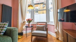 A cozy room with a large window, a chair, a small table with a vase of flowers, and a television on a stand. - Cute Cosy Cottage | Ramsgate Historic Area | 2bd (Kent)
